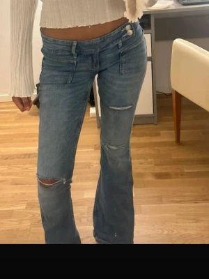 Blå slitna bootcut jeans - Säljer ett par ljusblå bootcut jeans med slitningar på knäna och låret. Jeansen har låg midja, klassiska fem fickor och stängs med två knappar framtill. Perfekta för en avslappnad och trendig look.