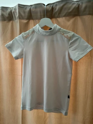 Vit topp med spets och pärlor - Snygg vit topp från CRW med korta ärmar, spetsdetaljer på axlarna och pärlor vid halsen. Toppen har en hög krage och är perfekt för dig som gillar en stilren men ändå unik look.