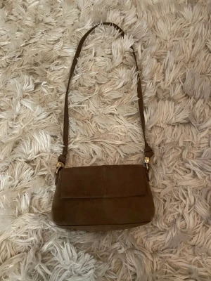 Brun axelväska i mocka - Snygg brun axelväska i mjuk mocka med enkel, rektangulär form och guldfärgade metalldetaljer vid axelremmen. Perfekt storlek för det viktigaste och ger en stilren vintagekänsla. 