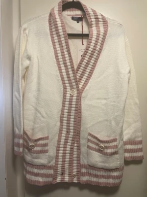 Vit kofta med rosa ränder från Felix Hardy - Lång, stickad kofta i vitt med breda rosa ränder längs krage, ärmslut och nederkant. Två fickor framtill med dekorativa knappar och randiga detaljer. 