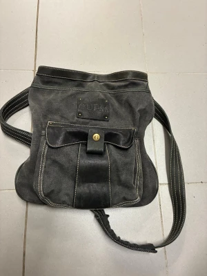 Svart axelväska från Guess - Cool svart axelväska från Guess i canvas med detaljer i läder och kontrastsömmar. Väskan har en stor ficka framtill med lock och guldfärgad knapp med G-logga. Perfekt för dig som vill ha en stilren och rymlig väska med streetkänsla.