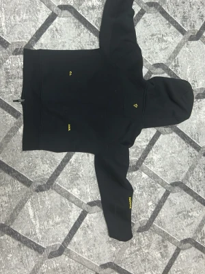 Svart Nike hoodie med dragkedja - Svart hoodie från Nike med hel dragkedja och två fickor framtill med svarta dragkedjor. Liten gul Nike-logga på bröstet och gula detaljer på ärmen. Hoodien har en klassisk huva och är tillverkad i ett mjukt, sportigt material.