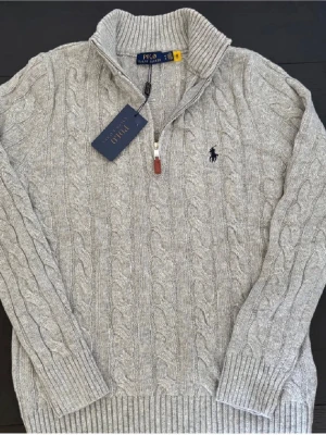 Polo Ralph Lauren halvzip - Kabelstickad grå tröja från Polo Ralph Lauren med halv dragkedja och hög krage. Klassisk broderad logga på bröstet och ribbade muddar. Tröjan är i nytt skick och på så sätt inga defekter. Hör av er vid frågor. 