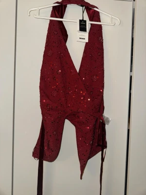 Vinröd omlottopp med paljetter Bik Bok - Vinröd omlottopp från Bik Bok med blommiga broderier och paljetter. Toppen har knyt i midjan och är ärmlös med djup v-ringning. Öppet rygg