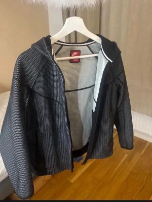 Nike tech - Snygg svartvit vindjacka från Nike med diskret rutmönster, hel dragkedja och huva. Jackan har två sidofickor med dragkedja och är tillverkad i ett lätt, vindtätt material som passar perfekt för blåsiga dagar. Sportig och stilren design.