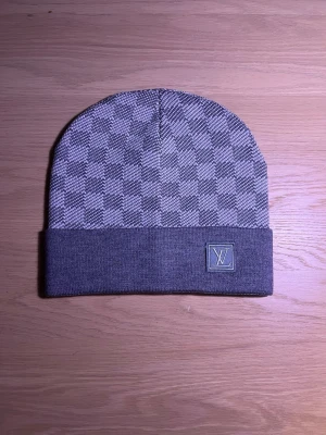 Grå Louis Vuitton mössa med rutmönster - Säljer en snygg grå mössa från Louis Vuitton med klassiskt rutmönster och vikt kant. Mössan har en diskret LV-logga framtill och är tillverkad i mjukt material som känns lyxigt och bekvämt.
