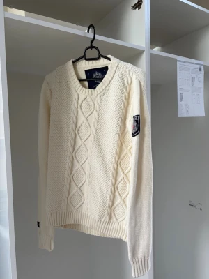 Vit kabelstickad tröja från Morris - Kabelstickad vit tröja från Morris med rund halsringning och snyggt mönster framtill. Tröjan har ett broderat märke på ärmen med brittisk flagga och ribbade muddar vid ärmslut och nederkant. Perfekt för en stilren och klassisk look.