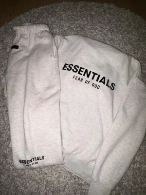 Essentials Fear of God hoodie grå - Ljusgrå hoodie från Essentials Fear of God med svart logga tryckt på ryggen och bröstet. Klassisk passform med ribbade muddar och midja. Tillverkad i mjukt bomullsmaterial som är perfekt för en avslappnad streetwear-look.