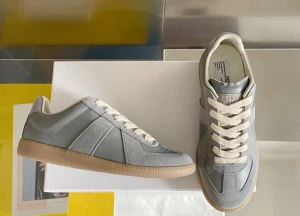 Grå sneakers från Maison Margiela - Stilrena sneakers i grått läder och mocka med vita snören och beige sula. Klassisk low-top design med diskreta paneler och ikonisk Margiela-etikett på plösen. Perfekta för en clean och modern look.