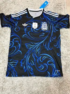 Argentina Bortaställ - Snygg Argentina fotbollströja från Adidas i svart med blått och turkost mönster. Tröjan har vita detaljer, tre ränder på axlarna och officiella AFA- och FIFA-märken på bröstet. Tillverkad i ett lätt och funktionellt material, perfekt för träning eller match.