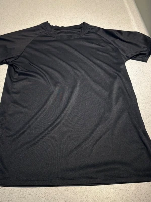 Svart sportig t-shirt i polyester - En enkel och stilren svart t-shirt i polyester med korta ärmar. Perfekt för träning eller chill dagar. Materialet är lätt och andas bra, vilket gör den skön att bära under aktivitet.