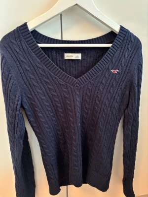 Mörkblå kabelstickad tröja Hollister - Mörkblå kabelstickad tröja från Hollister med V-ringning och lång ärm. Tröjan har ribbade muddar och ett litet broderat Hollister-logo på bröstet. Materialet är mjukt och följsamt, perfekt för kyligare dagar.
