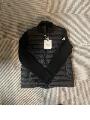 Svart cardigan från Moncler - Snygg svart Cardigan från Moncler med quiltad front och rygg samt stickade ärmar. Jackan har hög krage, två dragkedjeförsedda fickor och Moncler-logga på ärmen. Perfekt för kalla dagar och ger en stilren look.