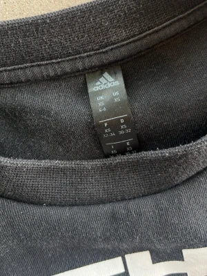 Svart sweatshirt från Adidas XS - Cool svart sweatshirt från Adidas med trycket 'THE THREE STRIPES' i vitt framtill. Tröjan har rund halsringning, långa ärmar och ribbade muddar vid ärmslut och nederkant. Perfekt för en sportig och avslappnad stil.