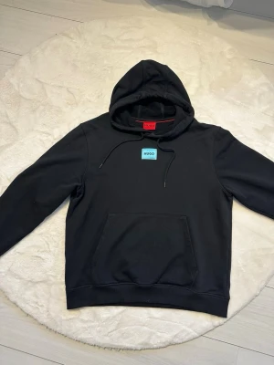 Svart hoodie från Hugo Boss - Svart hoodie från Hugo med klassisk huva och dragsnören. På bröstet sitter en liten blå patch med HUGO-loggan. Hoodien har en stor magficka och ribbade muddar vid ärmslut och nederkant. Perfekt för en avslappnad och stilren look.
