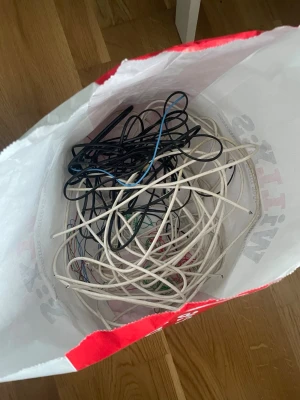 Påse med blandade elektroniksladdar -  En påse fylld med olika elektroniksladdar i färgerna svart, vit, röd, grön och blå. Sladdarna är av plast och har olika längder och tjocklekar, vissa med synliga koppartrådar. Perfekt för dig som gillar att pyssla med koppar och vill sälja det