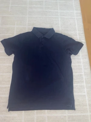 Mörkblå pikétröja med krage - En klassisk mörkblå pikétröja med krage och knappar framtill. Kortärmad modell i mjukt material, perfekt för en avslappnad och stilren look. Passar till jeans eller shorts för en enkel outfit.