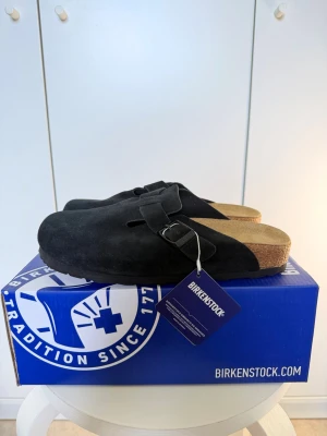 Svarta Birkenstock Boston tofflor - Säljer ett par svarta Birkenstock Boston tofflor i mocka med klassisk korksula och justerbart spänne. Tofflorna har en stilren och tidlös design som passar perfekt till både jeans och mjukisbyxor. Superbekväma och lätta att ta på sig.