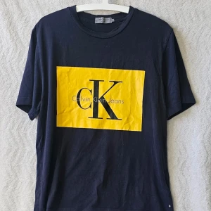 Svart Calvin Klein Jeans t-shirt - Snygg svart t-shirt från Calvin Klein Jeans med stort gult tryck och CK-logga på bröstet. Klassisk rund hals och korta ärmar. Tillverkad i mjuk bomull som känns skön mot huden. Perfekt för dig som gillar stilrena och trendiga plagg.