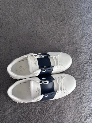 Valentino vita sneakers med blå detalj - Snygga vita sneakers från Valentino med bred mörkblå rem över mitten och klassiska vita snören. Skorna har en låg profil och coola nitar baktill på sulan. Tillverkade i läder för en lyxig känsla och stilren look.