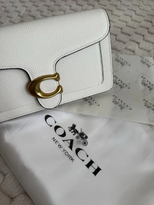 Vit handväska från Coach - Snygg vit handväska från Coach i strukturerat läder med guldfärgat C-spänne framtill. Väskan har en stilren och modern design med svarta kontrastsömmar och klassisk form. Perfekt accessoar för att lyfta din outfit.