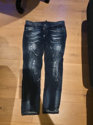 Mörkblå dsquered2 jeans  - Säljer ett par mörkblå jeans med slitningar och färgstänk för en cool, avslappnad look. Jeansen har raka ben, klassisk femficksmodell och detaljerade slitningar framtill. Perfekta för dig som gillar streetwear och vill ha en unik stil.