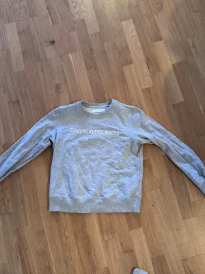 Grå sweatshirt från Calvin Klein Jeans - Snygg grå sweatshirt från Calvin Klein Jeans med vit logga tryckt över bröstet. Tröjan har rund halsringning, långa ärmar och ribbade muddar vid ärmslut och nederkant. Perfekt för en avslappnad och stilren look.