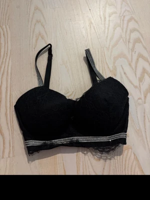 Svart spets-bh med strassdetalj Victorias secret - Snygg svart bh med vadderade kupor och spetsdetaljer från Victorias secret. Justerbara axelband och hakknäppning bak. Under bysten sitter en rad med små strass-stenar som ger extra bling. Perfekt för dig som gillar en mix av spets och glam.