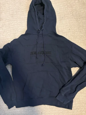 Mörkblå hoodie från Zadig & Voltaire - Snygg mörkblå hoodie från Zadig & Voltaire med tryck på bröstet och stor känguruficka framtill. Klassisk huva med dragsko och ribbade muddar vid ärmar och nederkant. Perfekt för en avslappnad och trendig look.