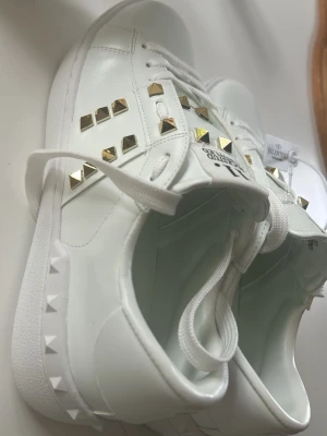 Vita Valentino Rockstud sneakers - Snygga vita sneakers från Valentino med guldiga nitar i Rockstud-stil. Skorna har klassisk låg profil, vita skosnören och är tillverkade i slätt läder. Perfekta för dig som vill ha en lyxig och trendig look med extra edge.
