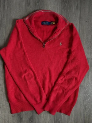 Röd halvzip tröja från Polo Ralph Lauren - Snygg röd tröja från Polo Ralph Lauren med halv dragkedja och klassisk broderad logga på bröstet. Tröjan har ribbade muddar och krage, perfekt för lager på lager. Materialet är mjukt och bekvämt, passar till många olika stilar.