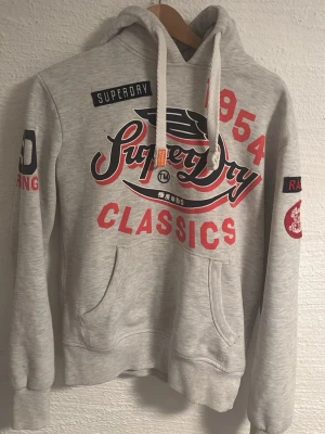 Superdry grå hoodie med tryck - Snygg grå hoodie från Superdry med stora röda och svarta tryck på bröstet och armarna. Klassisk känguruficka framtill, dragsko i huvan och broderade detaljer. Perfekt för en avslappnad streetstyle-look.