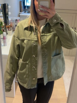 Grön Zara jacka - Snygg overshirt i ljusgrönt denimliknande material med stora framfickor och silverfärgade knappar. Rak passform och klassisk krage ger en avslappnad vibe. Perfekt att slänga över en hoodie eller t-shirt för en cool lager-på-lager-look.