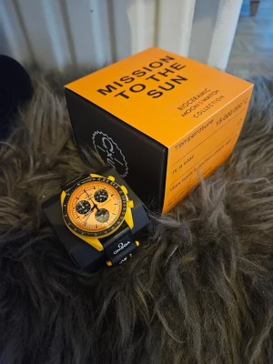 Omega x Swatch Mission to the Sun Remix - Snygg Omega x Swatch Speedmaster Mission to the Sun med gul boett och svart urtavla. Klockan har kronograf, tachymeter och svart textilband med Omega-logga. Perfekt för dig som gillar unika och färgstarka accessoarer.