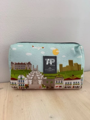 Necessär TAP Portugal x Castelbel - Necessär med färgglatt tryck av stadsmotiv, flygplan och sol på ljusblå bakgrund. Märkt med TAP Portugal och Castelbel. Praktisk dragkedja upptill och rektangulär form. Perfekt för småprylar eller smink.