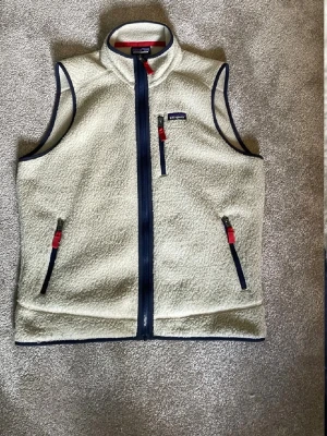 Vit fleeceväst från Patagonia - Snygg vit fleeceväst från Patagonia med marinblå detaljer och röda dragkedjor. Västen har hög krage, tre fickor med dragkedja och klassisk Patagonia-logga på bröstet. Perfekt lager-på-lager-plagg för kyliga dagar.