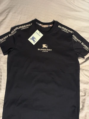 Svart Burberry t-shirt med logga - Snygg svart t-shirt från Burberry med klassisk logga och text på bröstet samt axlarna. T-shirten har rund hals och korta ärmar. Tillverkad i mjuk bomull för skön känsla. Perfekt för dig som vill ha en stilren och exklusiv look.