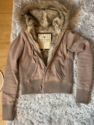 Beige teddyjacka från Abercrombie & Fitch - Mysig beige teddyjacka från Abercrombie & Fitch med fuskpälsfoder i huvan och insidan. Jackan har dragkedja framtill, ribbade muddar och två fickor. Perfekt för kyliga dagar och riktigt snygg till jeans eller leggings. Passar både S och Xs🥰