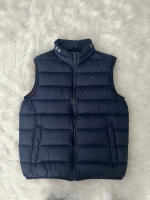 Mörkblå dunväst Tommy Hilfiger - Snygg mörkblå dunväst från Tommy Hilfiger med hög krage där loggan är broderad. Västen har två fickor med dragkedja och diskret logga på bröstet. Klassisk quiltad design och lätt vadderad för extra värme. Perfekt för lager-på-lager-stil.