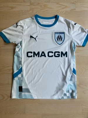 Marseille matchtröja Puma vit/blå - Säljer en vit matchtröja från Puma för Olympique de Marseille med blå detaljer och klubbmärke på bröstet. Tröjan har korta ärmar, rund hals och tryckt sponsorlogga framtill. Materialet är lätt och andas, perfekt för fotbollsträning.