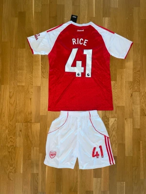Declan Rice Arsenal matchställ #41 - Snyggt Arsenal matchställ med Rice #41. Tröjan är röd med vita ärmar och detaljer, och har klubbmärke samt sponsortryck framtill. Shortsen är vita med röda detaljer och klubbmärke. Perfekt för fotbollsträning eller att bära på matchdag.