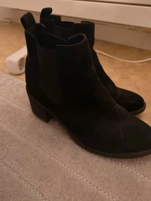 Svarta Chelsea boots med klack - Säljer ett par svarta Chelsea boots i mocka-look med bred resår på sidorna och stadig blockklack. Klassisk siluett som är enkel att matcha till olika outfits. Perfekta för dig som vill ha både stil och komfort. Inga typer av skador men är i använt skick. Lite smutsig men som går att tvätta