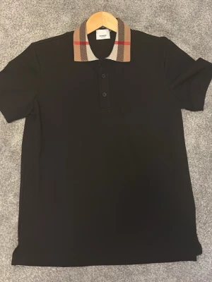 Svart pikétröja med rutig krage Burberry - Svart pikétröja från Burberry med klassisk rutig krage i beige, rött och svart. Tröjan har korta ärmar och knäppning framtill med tre knappar. Perfekt för en stilren och trendig look.