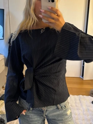 Mörkblå randig blus med knytband - Snygg mörkblå blus med tunna vita ränder och breda långa ärmar. Blusen har ett knytband som frmhäver midjan.