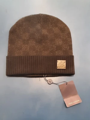 Brun mössa från Louis Vuitton - Snygg brun mössa från Louis Vuitton med diskret mönster och ribbad uppvikt kant. Mössan har en metallbricka med Louis Vuitton-logga framtill och är tillverkad i ett mjukt, stickat material. Perfekt accessoar för att lyfta din stil.