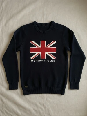 Morris R. Club stickad tröja - Snygg mörkblå stickad tröja från Morris R. Club med stor Union Jack-flagga i rött, vitt och blått på bröstet. Tröjan har rund halsringning och ribbade muddar vid ärmslut och nederkant. Perfekt för dig som gillar preppy stil. Archive stil jag är 173 och passar helt okej tror folk som är 175 också hade kunnat passa i den bra