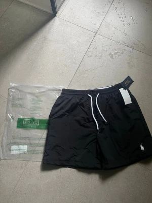 Svarta badbyxor från Polo Ralph Lauren - Snygga svarta badbyxor från Polo Ralph Lauren med vit dragsko i midjan och klassisk logga broderad på benet. Tillverkade i snabbtorkande material och har en avslappnad passform. Perfekta för stranden eller poolen. Passar M!
