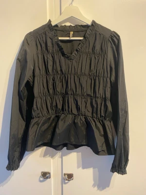 Svart volangblus med v-ringning - Snygg svart blus med v-ringning och volang nedtill. Blusen har långa ärmar med resår vid ärmslut och smockdetaljer över hela framsidan. 