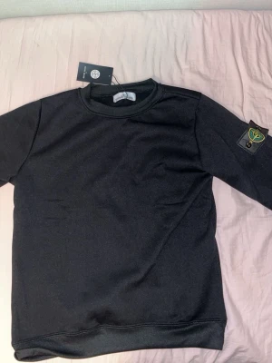 Svart sweatshirt från Stone Island - Säljer en svart sweatshirt från Stone Island med klassisk rund hals och ikonisk logotyp-patch på vänster ärm. Säljer tröjan för råkade beställa fel storlek men annars är tröjan helt ny och oanvänd. 