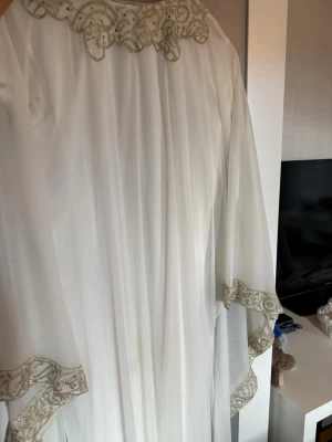 Vit långklänning med broderade detaljer - Elegant vit långklänning med vackra broderade detaljer i guld runt halsringning och ärmslut. Klänningen har en lös passform och vida ärmar som ger ett flytande intryck. Perfekt för dig som vill ha en unik och stilren look.